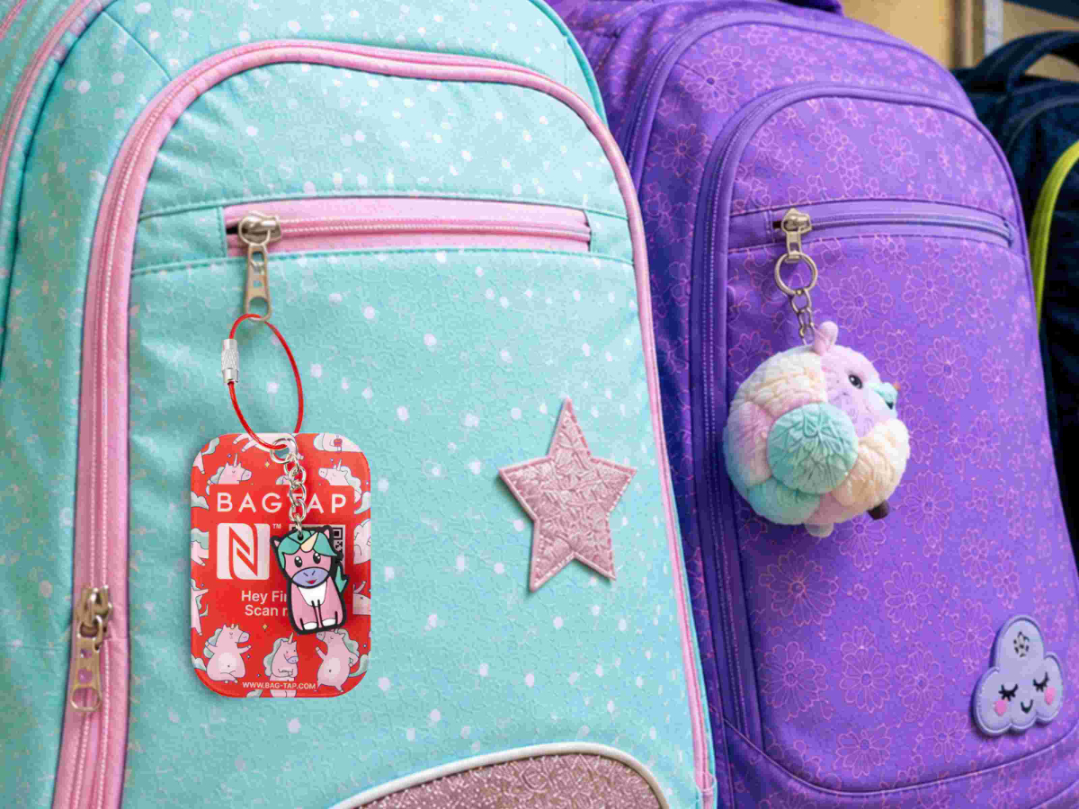 Ein türkisfarbener Kinder-Rucksack mit einem roten BAGTAP-Adressanhänger im Einhorn-Design, daneben steht ein lila Rucksack.